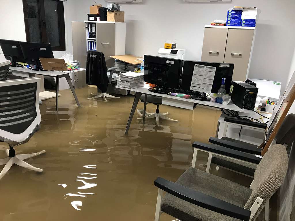 Officeflooding Office space affected by flooding with water covering the floor, desks, computers, and chairs, showing the need for professional water damage restoration services.